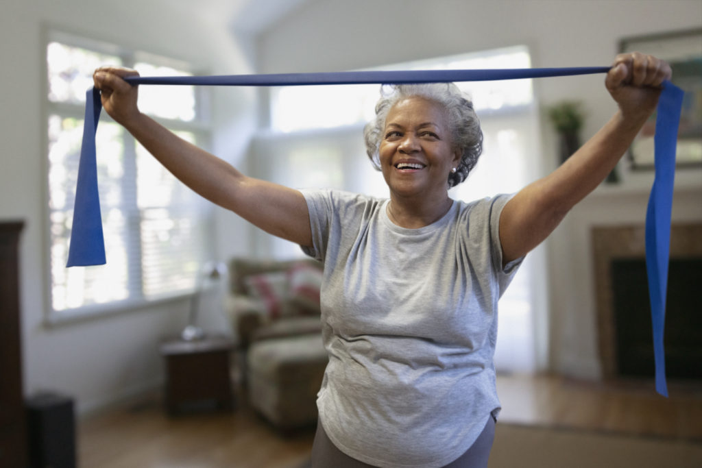Image shows active ageing through and older black woman in a grey t-shirt using a blue resistance band with both hands above her head. 