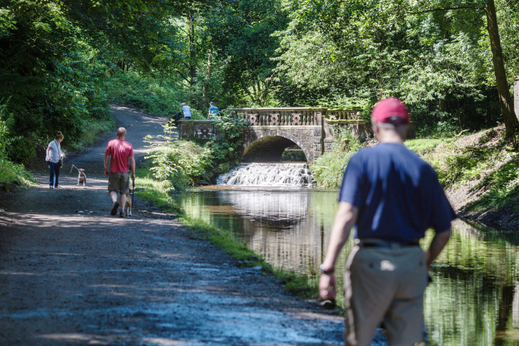 Image highlights active living by a man in a blue t-shirt, red cap and khaki trousers walking on a path by the side of a river in a forest. A bridge with a waterfall is in the background with more walkers. 