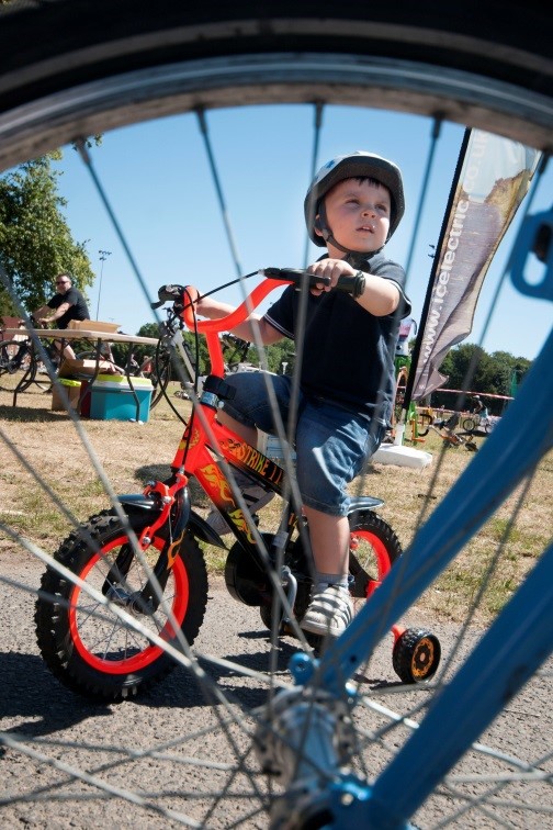 Image shows a young, white boy on a red bike with stabilisers wearing a helmet, black t-shirt and denim shorts. This photo was taken through the spindles of another bike. In a park which links to the active start article.