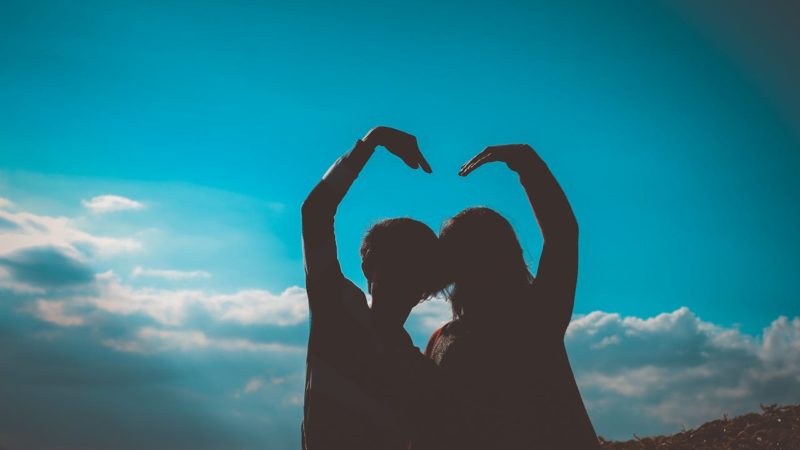 couple making a heart sign with their arms. The couple are in shadow against a blue sky.